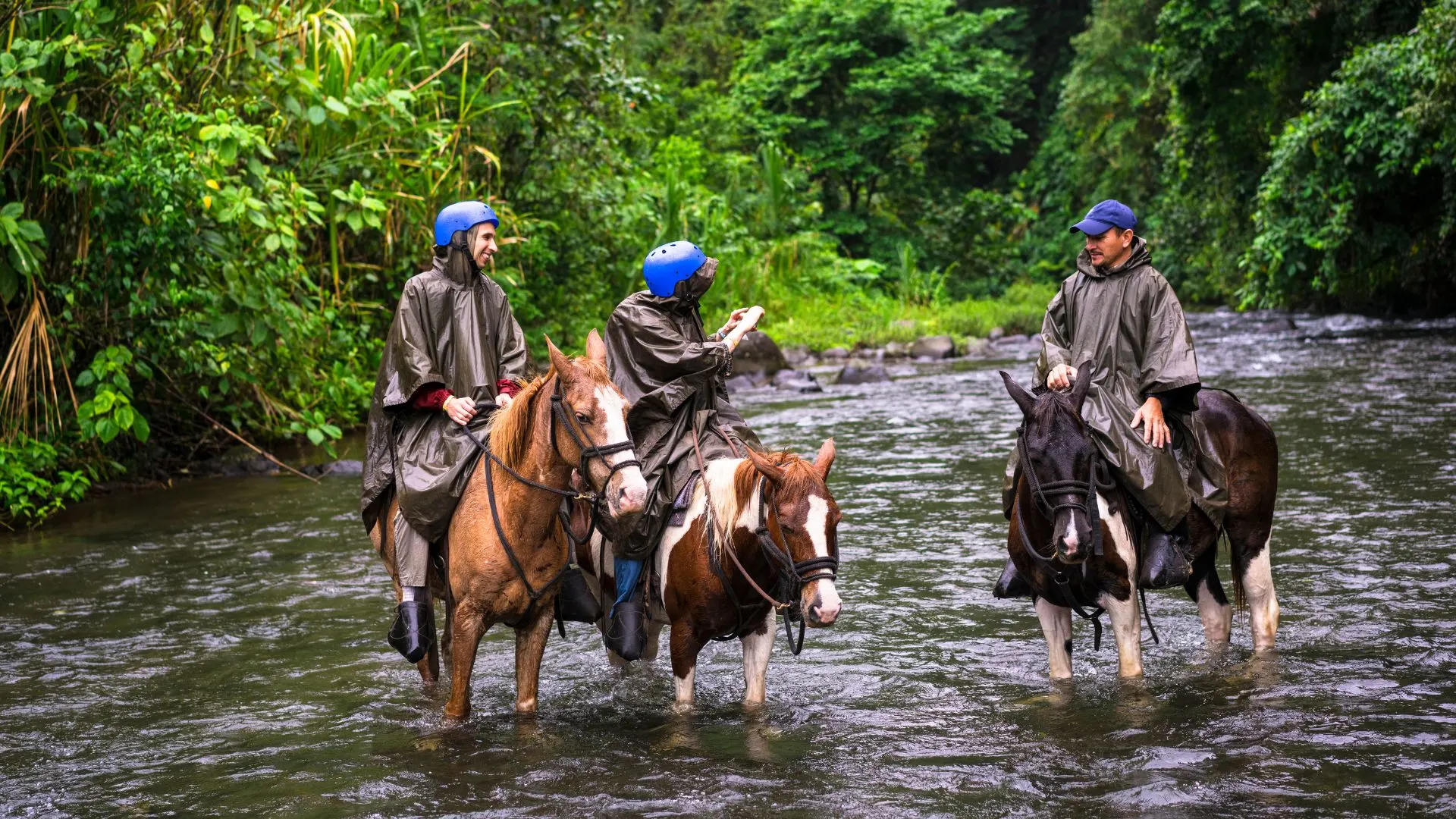 Three people on horses wearing rain ponchos and helmets traverse a shallow river in a lush forest, experiencing the thrill of Costa Rican tours.